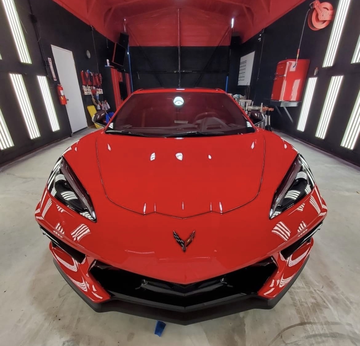 A red Corvette head-on in the detail bay, LED panels reflected in the hood